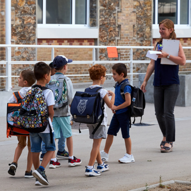 Une institutrice dirige un rang de cinq élèves avec des cartables à travers la cours de l'école.